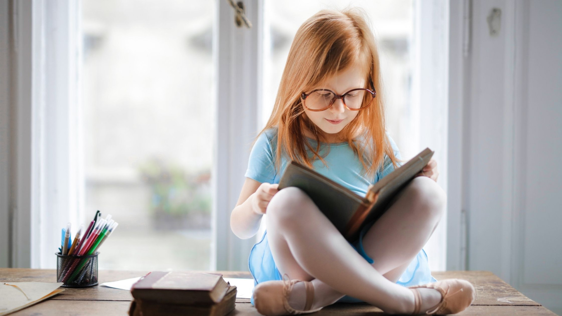 Small girl wearing glasses sitting in a sunny window reading a book
